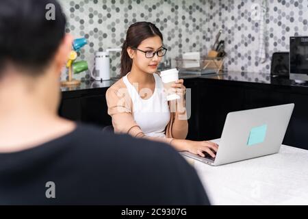 Asiatische Frau Arbeit von zu Hause in der Küche trinken Kaffee mitnehmen, während Mann in der Ferne im Vordergrund arbeiten, während Stadt Sperre von Coronavirus covid-19 pa Stockfoto