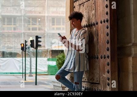 Der junge Afro lehnt sich an ein hölzernes Tor und schaut auf seine Telefon Stockfoto