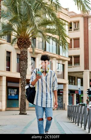 Ein junger afrohaariger Mann geht die Straße hinunter und schaut zu Sein Handy Stockfoto