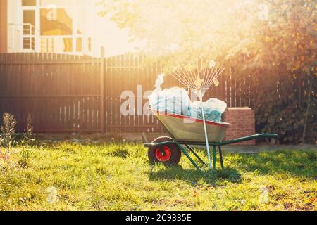 Garten Schubkarre mit Müllsäcke und Rechen stehen auf dem Gras in der Nähe des Zauns in der warmen Glow. Stockfoto