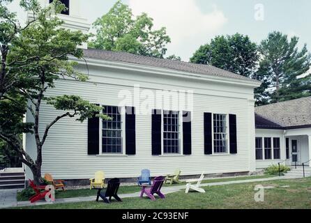 Bunte Adirondack Stühle sitzen leer, sechs Meter auseinander für soziale Distanzierung außerhalb einer weißen New England Kirche in der Sommersonne. Die Distanzierung Stockfoto