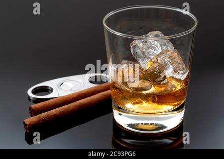 Tabakrauchen und Alkoholtrinken Konzept mit Glas Whiskey auf den Felsen, Zigarren, Zigarrenschneider aus Metall und Eis auf dunklem Hintergrund Stockfoto