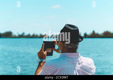 Älterer Tourist, der ein Foto mit seinem Smartphone gemacht hat und eine Einweg-Gesichtsmaske trägt. Stockfoto