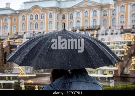 St. Petersburg, Russland - 23. Juli 2020 EIN Paar steht unter einem Schirm und schaut im strömenden Regen auf den Großen Kaskadenbrunnen und die Sehenswürdigkeiten des Königspalastensembles in Peterhof, Russland Stockfoto