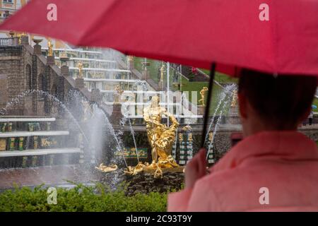 St. Petersburg, Russland - 23. Juli 2020 EINE Frau steht unter einem Regenschirm und schaut bei strömendem Regen auf den Großen Kaskadenbrunnen und die Sehenswürdigkeiten des Königspalastensembles in Peterhof, Russland Stockfoto