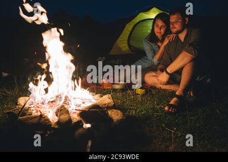 Lächelndes fröhliches Paar, das vor einem kochenden Abendessen sitzt. Camping-Konzept Stockfoto