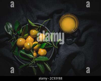 Mandarinen mit grünen Blättern in blauer Schale und Glas frischen Mandarinensaft auf dunklem Hintergrund. Overhead-Aufnahme. Stockfoto