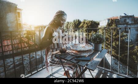 Eine hinreißende junge schwarze Frau sitzt mit einem Glas Wein zu Mittag und schneidet eine leckere Pizza in Stücke Stockfoto