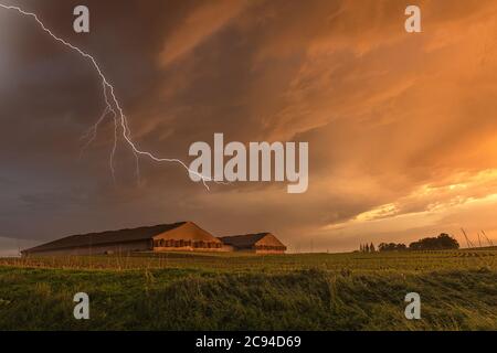 Ein Gewitter bei Sonnenuntergang passiert eine Bauerngemeinschaft mit einem Raupenwagen, der gegen den orangefarbenen, dunklen Himmel gerahmt ist. Stockfoto