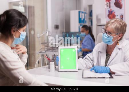 Arzt mit Gesichtsmaske Blick auf Tablette mit grünem Bildschirm während der Konsultation mit kranken Patienten. Krankenschwester trägt blaue Uniform. Stockfoto
