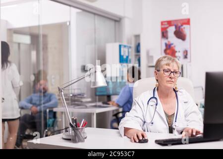 Ältere Frau Arzt arbeitet am Computer im Krankenhaus Büro, während junge Sanitäter mit behinderten Patienten im Rollstuhl auf Klinikkorridor und Krankenschwester in blauer Uniform diskutiert. Stockfoto