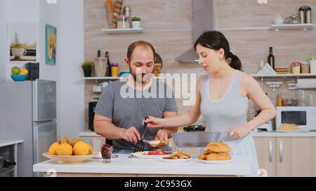 Frau kocht Eier für den Mann während des Frühstücks, während er Butter auf geröstetem Brot schmiert. Tragen Pyjamas am Morgen, Vorbereitung Mahlzeit zusammen, junge glückliche Paar Liebe und Heiratsege Stockfoto