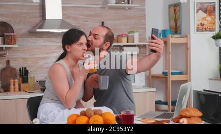 Glückliches junges Paar, das beim Frühstück Selfies macht. Fröhliche verheiratete Mann und Frau machen lustige Gesichter, während ein Foto während des Frühstücks in der Küche. Stockfoto