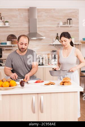 Frau kocht Eier für den Mann während des Frühstücks, während er Butter auf geröstetem Brot schmiert. Tragen Pyjamas am Morgen, Vorbereitung Mahlzeit zusammen, junge glückliche Paar Liebe und Heiratsege Stockfoto