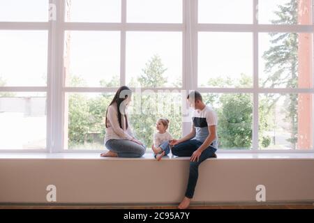 Kleines Mädchen mit Mama und Papa sitzt am großen Fenster im Studio. Familienphotosession Stockfoto