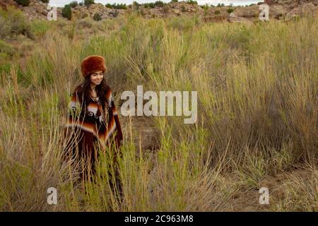 Junge hispanische Frau, gekleidet als Trapper in Poncho und Pelzhut, wandert mit hohem Gras durch die Wüstenlandschaft in der ojito Wilderness, New Mexico Stockfoto