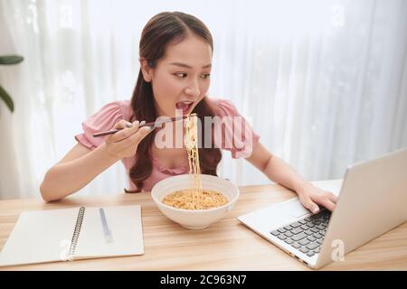 Junge asiatische Frau essen Nudeln und arbeiten mit Laptop zu Hause. Stockfoto