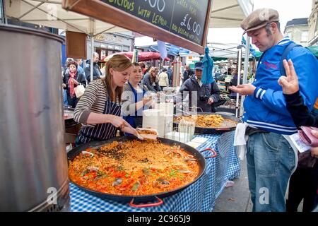 London, Großbritannien - Straßenszene auf dem Markt in der Portobello Road. Eine junge Marktfrau verkauft Paella an ihrem Marktstand. Der Markt in Notting Hi Stockfoto