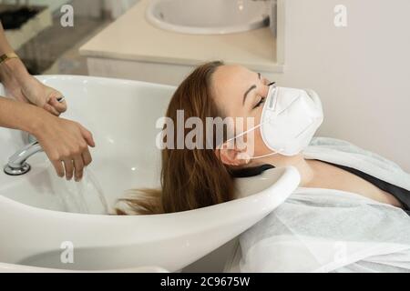 Junges Mädchen mit langen braunen Haaren waschen ihren Kopf bei der Friseurin Social Distanzing. Verwendung der Covid-19 Gesichtsmaske Stockfoto