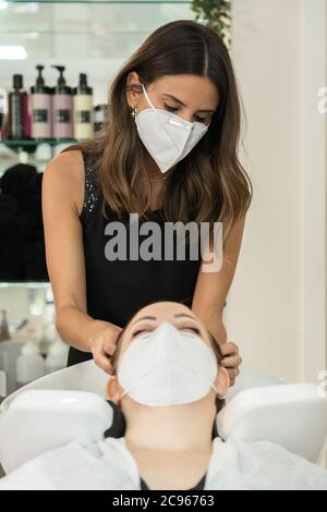 Ein hübscher Friseur, der einem jungen Kunden im Salon die Haare wäscht. Soziale Distanzierung. Verwendung der Covid-19 Gesichtsmaske. Vertikal Stockfoto