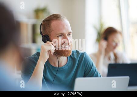 Reifer bärtiger Mann mit Kopfhörern, der am Tisch mit Laptop im Büro arbeitet Stockfoto