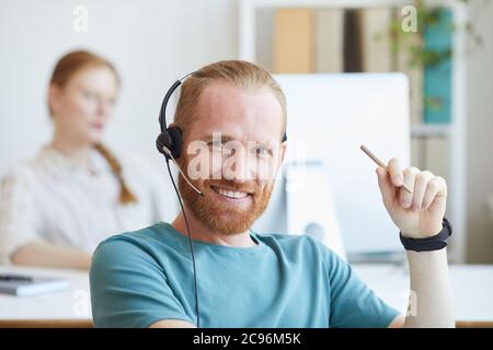 Porträt eines jungen bärtigen Mannes im Headset, der während der Arbeit im Büro die Kamera anlächelt Stockfoto