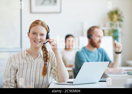 Porträt einer jungen Kundendienstfrau in Kopfhörern, die mit Laptop am Tisch sitzt und im Büro die Kamera anlächelt Stockfoto