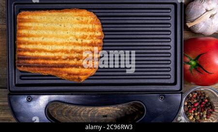 Gegrilltes weißes Toastbrot mit goldener Kruste in Streifen liegt in offener schwarzer Sandwich-Maker, auf Holz rustikalen Tisch. Gewürze, Tomaten, Knoblauch. Schnelles CO Stockfoto