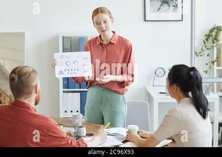 Junge Geschäftsfrau zeigt auf Geschäftsdiagramme in ihren Händen Sie gibt Geschäftspräsentation für ihre Kollegen im Büro Stockfoto