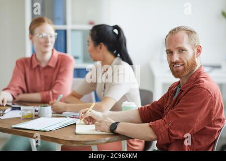 Porträt eines jungen bärtigen Geschäftsmannes, der während eines Geschäftstreffens am Tisch mit seinen Kollegen auf die Kamera schaut Stockfoto