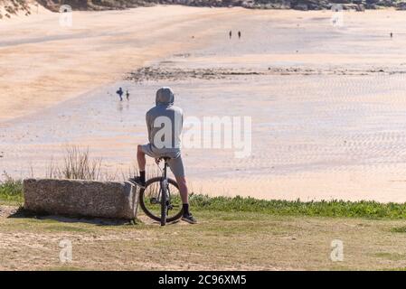Ein Mann mit Hoodie sitzt auf seinem Mountainbike mit Blick auf Fistral Beach in Newquay in Cornwall. Stockfoto