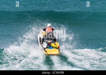 Spektakuläre Action als RNLI Rettungsschwimmer fährt einen Jetski über eine Welle bei Fistral in Newquay in Cornwall. Stockfoto