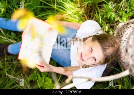 Teenager-Mädchen in weißem T-Shirt sitzen am Baum im Wald und lesen Interessantes Buch. Tolle Idee und Aktivität, um Sommerferien zu verbringen. Stockfoto