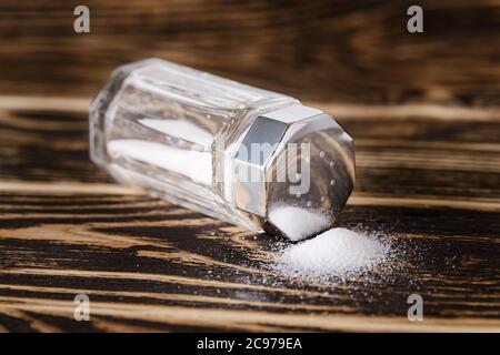 Aus dem Glas verschüttete Salzstreuer mit silbernem Metalldeckel liegt auf seiner Seite auf einem dunklen Holztisch. Low-Key-Fotografie. Vorderansicht. Stockfoto