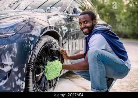 Seitenansicht des hübschen jungen afrikanischen Mann mit grünen Mikrofaser Auto Waschhandschuh für die Reinigung der Felgen seines modernen Autos im Freien. Luxus blaue Elektroauto in Stockfoto