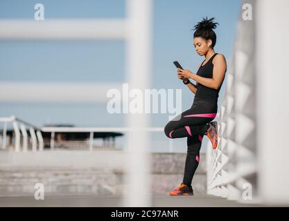 Sport-Blog und Outdoor-Workout. Serious african american Mädchen in Mode Sportbekleidung mit kabellosen Kopfhörern, Eingabe auf Smartphone Stockfoto