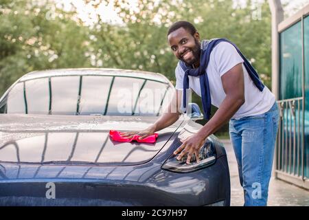 Junger glücklich lächelnder afrikanischer Mann, der seine futuristische Elektroauto-Haube und Scheinwerfer mit rotem Mikrofasertuch putzt und auf die Kamera schaut. Autowaschanlage im Freien Stockfoto