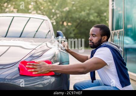 Junger afrikanischer Mann wascht und wischt seinen modernen blau grauen Autoscheinwerfer an der Outdoor-Autowaschanlage Self Service. Dunkelhäutiger Mann mit roter Mikrofaser Stockfoto