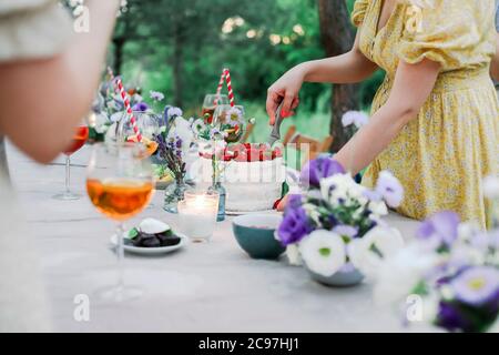 Nicht erkennbare Frau in Kleid schneiden süßen Kuchen auf Banketttisch während der Feiertagsfeier am Sommertag im Garten Stockfoto