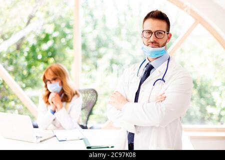 Schuss von männlichen Arzt trägt Gesichtsmaske, während im Büro des Arztes stehen. Im Hintergrund weibliche Ärztin, die am Laptop arbeitet. Stockfoto