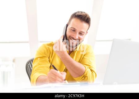 Aufnahme eines glücklichen Geschäftsmannes, der einen Anruf macht und etwas schreibt, während er hinter seinem Laptop am Schreibtisch sitzt. Stockfoto
