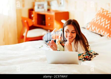 Youg weiblich in häuslicher Kleidung sitzt auf dem Bett mit einem Glas Wein und Surfen Netbook während zu Hause ausruhen Stockfoto