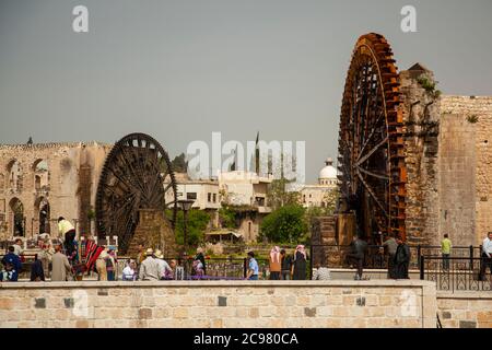 Hama/Syria 03/27/2010: Stadtbild der syrischen Stadt Hama, mit historischen Wasserrädern (Norias) im Hintergrund. Das Bild zeigt die Menschen vor Ort in traditi Stockfoto