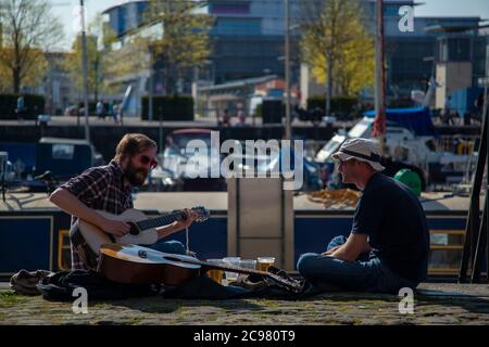 Bristol/UK 04/17/2010: Zwei Männer sitzen mit gekreuzten Beinen auf dem Boden, trinken Bier und spielen klassische Gitarre in der Hafenregion Bristol, UK ist einer bärtig Stockfoto
