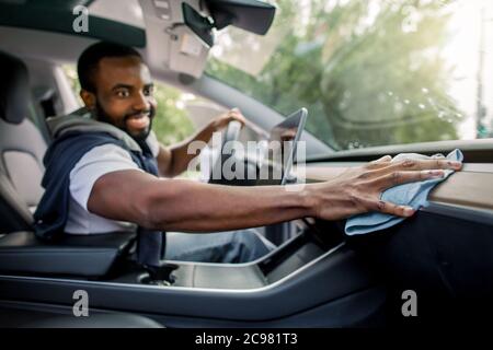 Junger gutaussehender lächelnder afrikanischer Mann, der die Fahrzeugkonsole und das Bedienfeld seines modernen Elektroautos mit Mikrofasertuch putzt. Autowäsche und Details Stockfoto