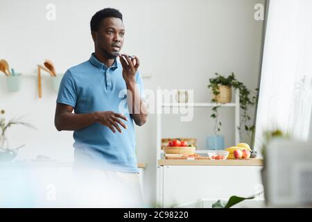 Afrikanischer junger Mann, der das Mobiltelefon in der Hand hält und Audiobotschaft aufzeichnet, während er zu Hause im Zimmer steht Stockfoto
