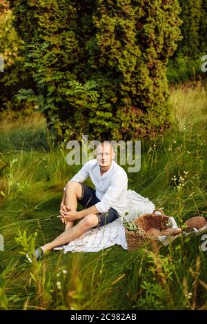 Schöner Mann, der im Park mit einem Korb auf einer Decke sitzt Stockfoto