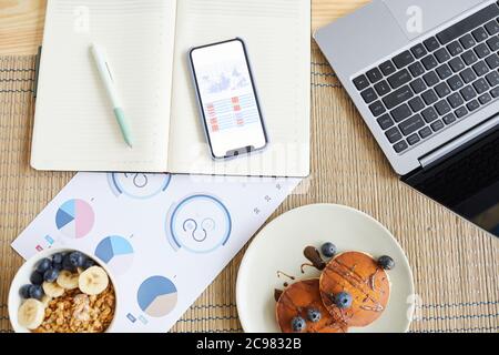 High-Angle-Ansicht von Geschäftsdokumenten mit Diagrammen und Grafiken mit Laptop und Frühstück auf dem Tisch zu Hause Stockfoto