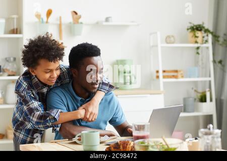 Afrikanischer Mann sitzt am Tisch und arbeitet auf Laptop mit seinem Sohn umarmt ihn sind sie in der Küche Stockfoto