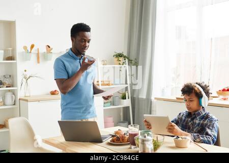 Afrikanischer junger Vater, der am Telefon spricht und Dokumente prüft, die er zu Hause arbeitet, während sein Sohn in der Küche Spiele auf einem digitalen Tablet spielt Stockfoto